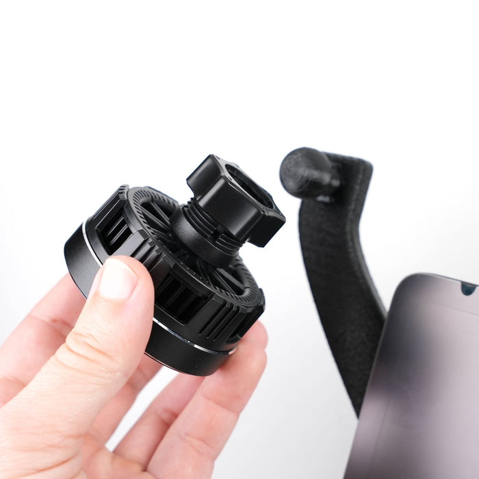 A hand holding a black phone mount with a gear mechanism, positioned next to a smartphone on a black stand.