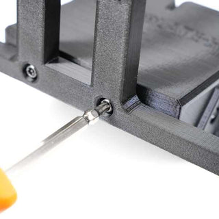 A screwdriver with an orange handle is inserted into a screw on a partially visible black plastic object, possibly an assembly or repair process.