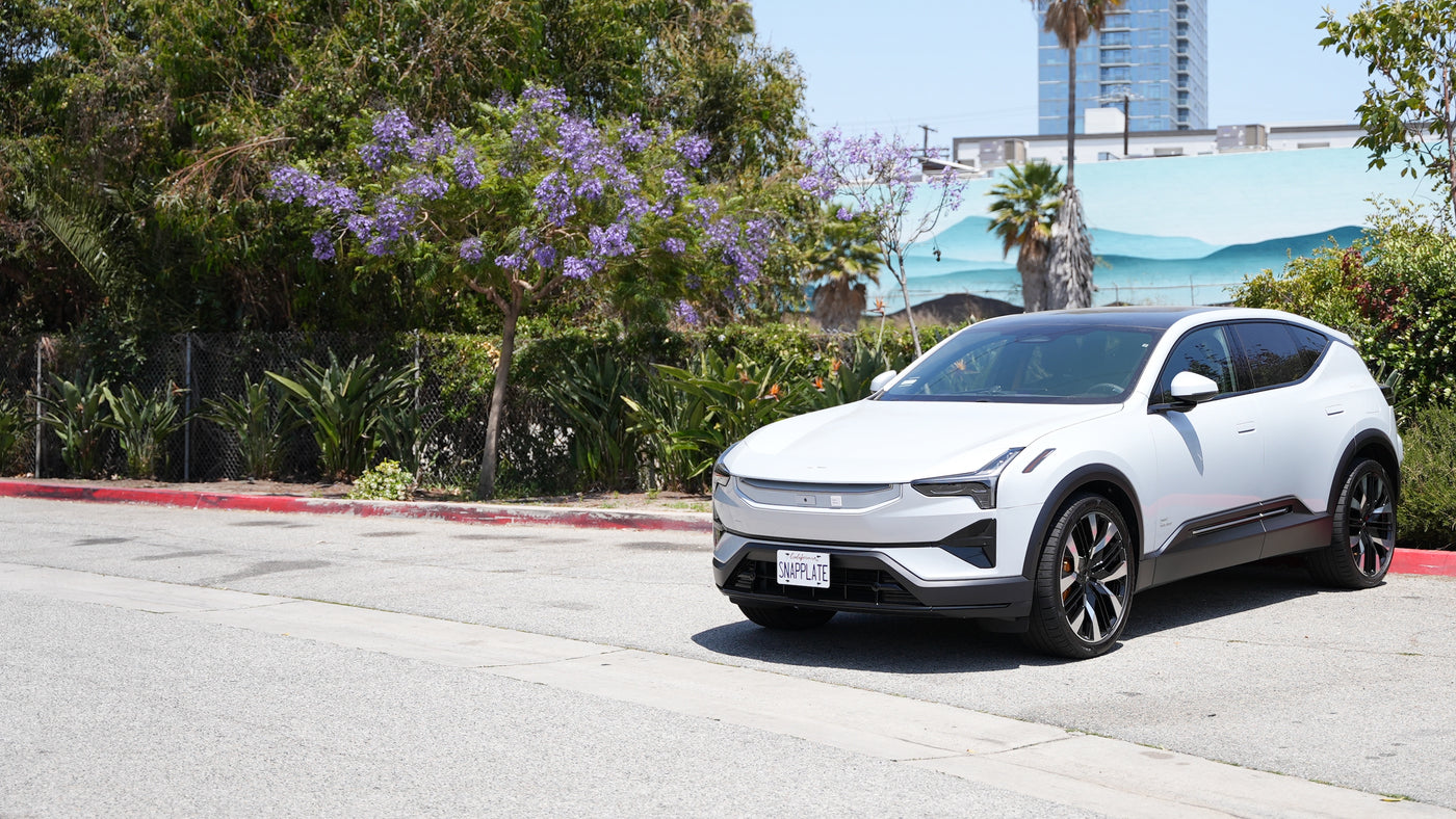 A white electric SUV is parked on the side of a street near trees, shrubs, and a mural, with a tall building visible in the background.