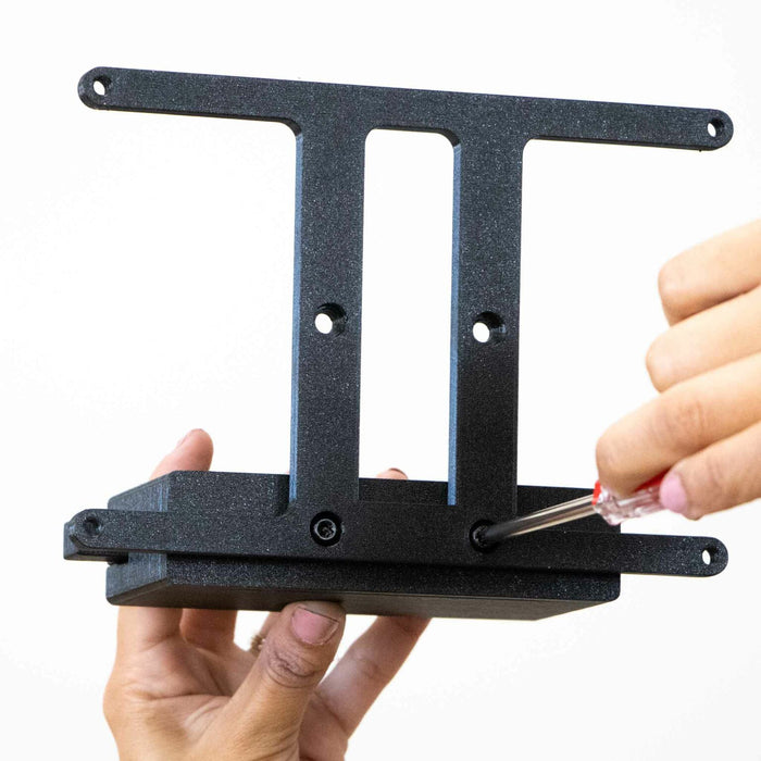 A person uses a screwdriver to adjust or assemble a black metal bracket with four mounting holes against a white background.
