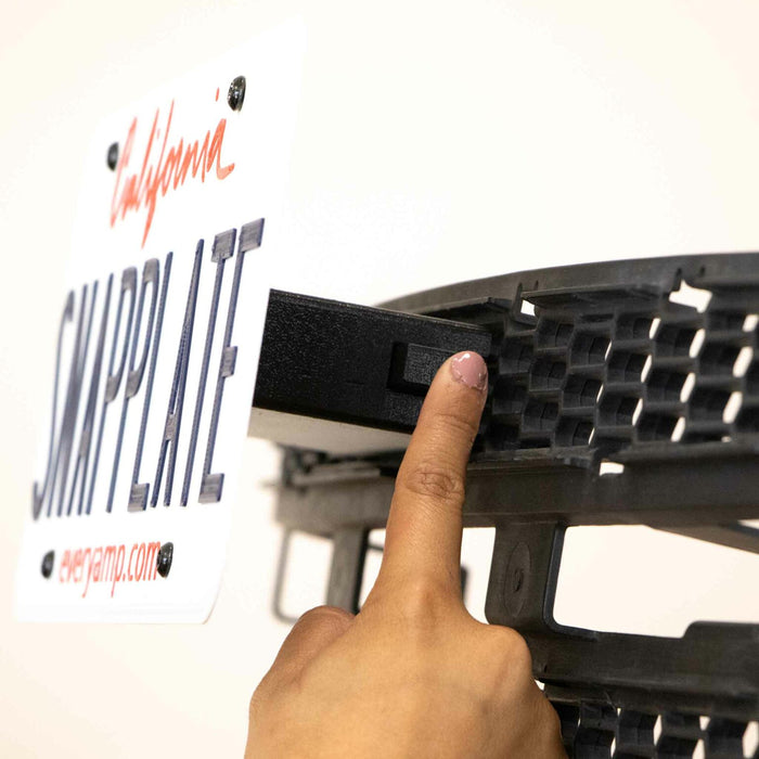 A hand pointing at a mounting bracket behind a California sample license plate on a vehicle bumper.