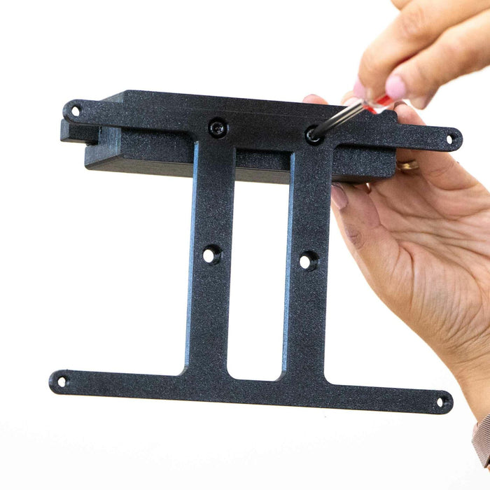 Hands using a screwdriver to assemble a black metal bracket with pre-drilled holes for mounting, against a white background.