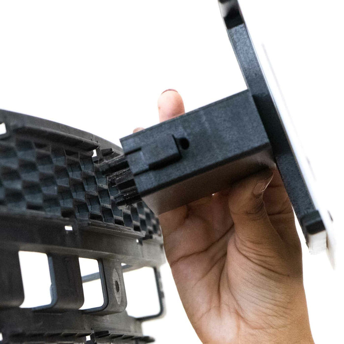 A person inserts a rectangular black bracket into a grille-like surface, holding the edge with their hand against a white background.