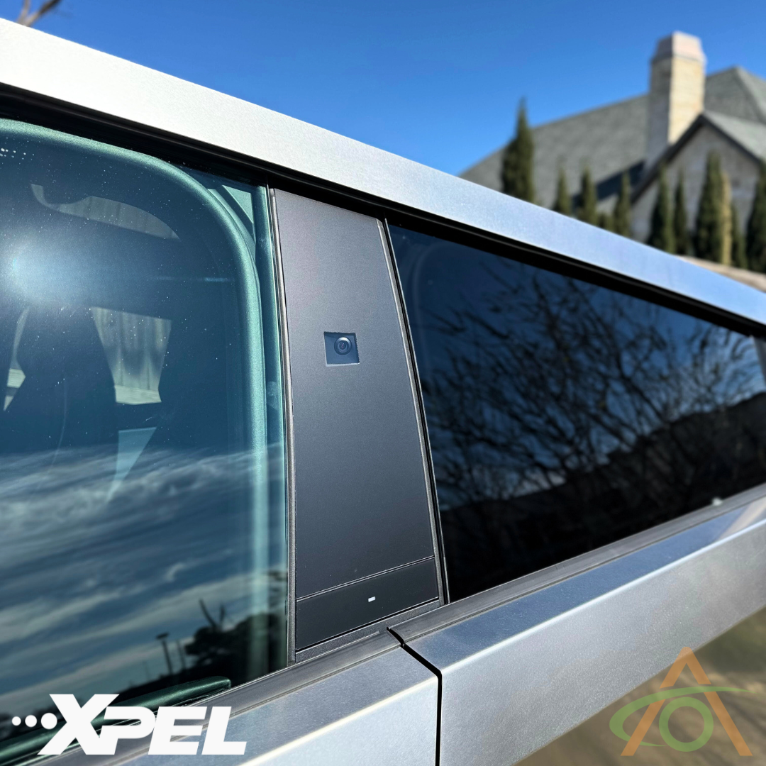 Close-up of a vehicle exterior featuring a camera on the pillar with reflective windows, protected by Abstract Oceans B & C Pillar Protection - PPF for Cybertruck. The branding includes XPEL and an orange logo, with a house and trees in the background emphasizing its appeal.
