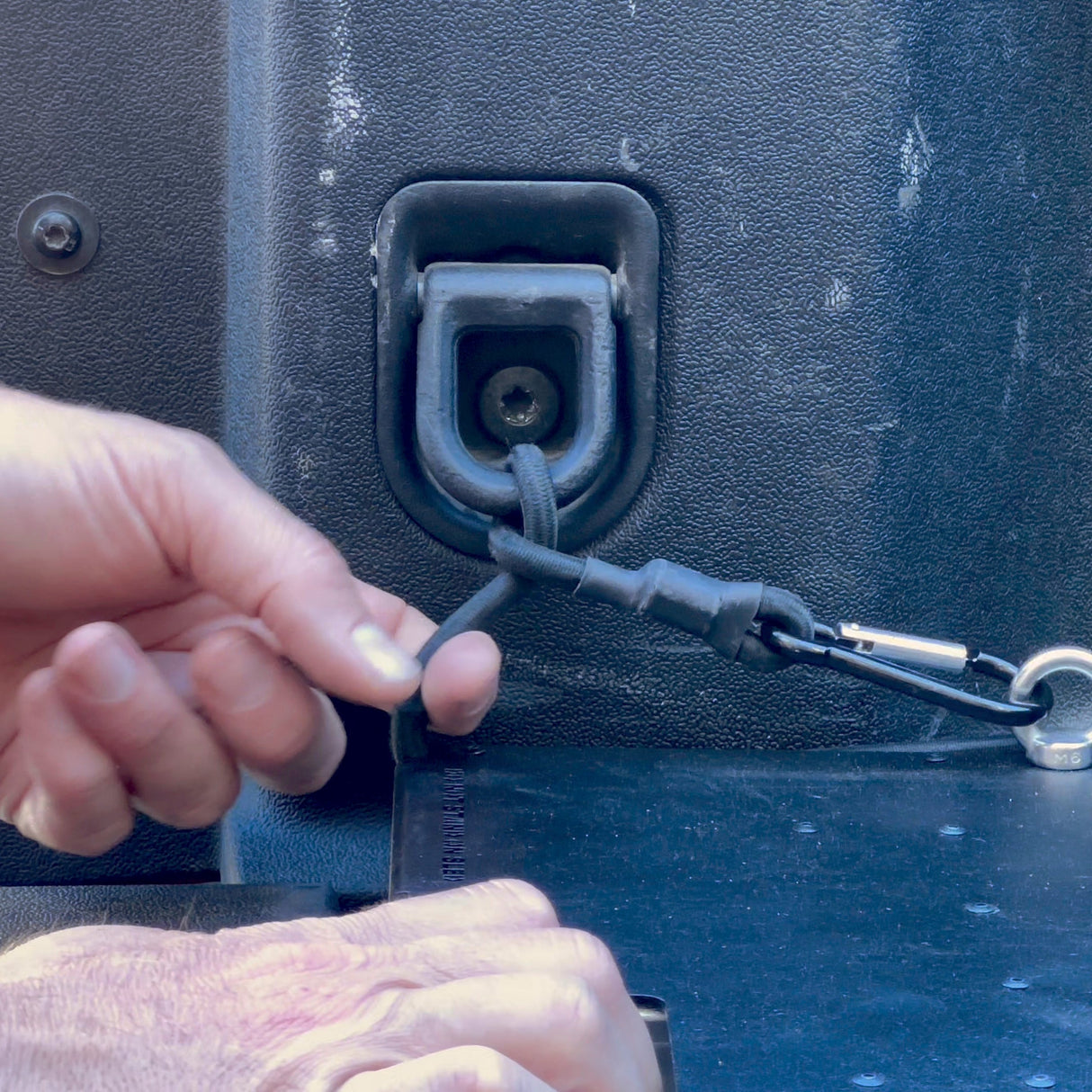 A close-up of hands attaching a black cord to a metal anchor inside a truck bed, highlighting textured surfaces and the Thunderbolt Adventure Supply R1T Bed Glider—perfect for maximizing truck bed storage.