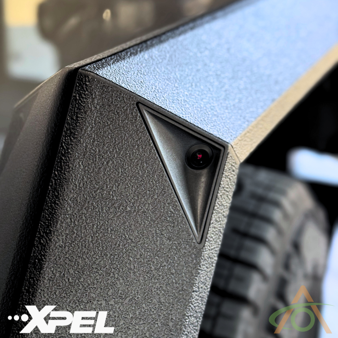 Close-up of a textured black Cybertruck fender flare featuring a triangular camera. The Abstract Ocean logo and Xpel hint at the durable protection of Ultra-Tough Xpel Armor film.