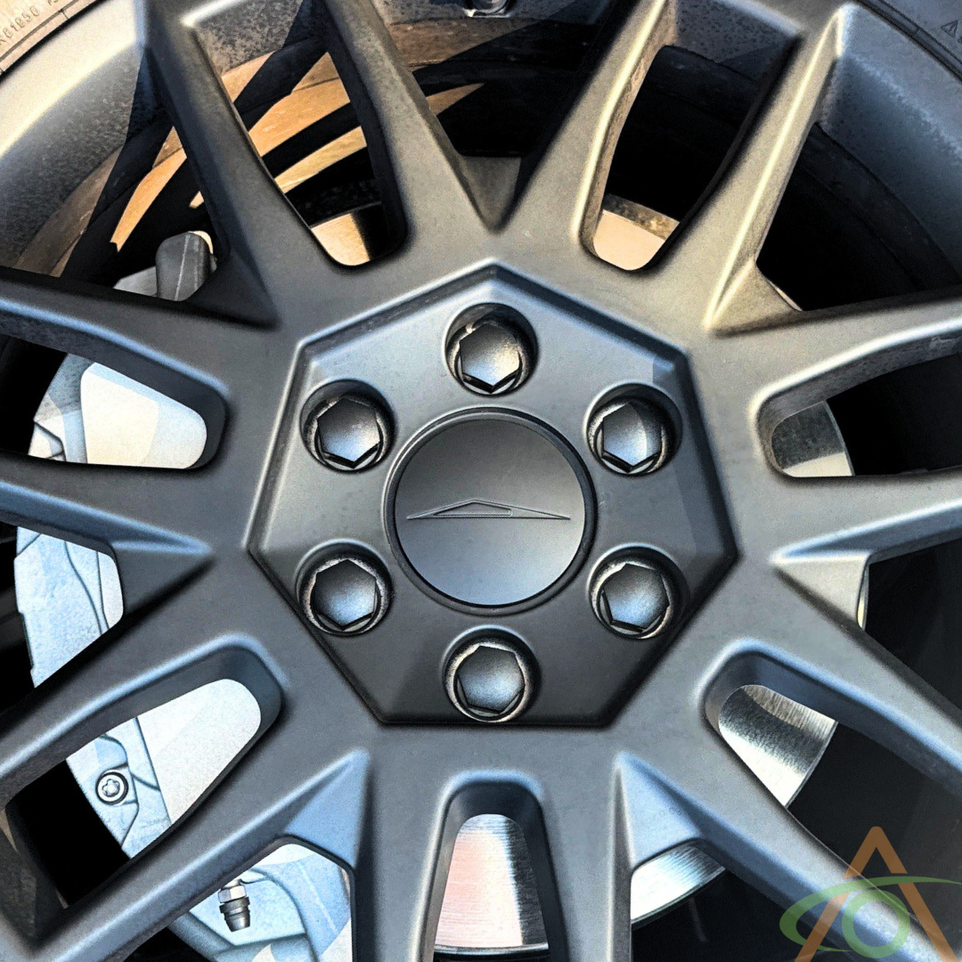 A close-up of a dark alloy car wheel with a geometric design features five lug nuts arranged in a star pattern. The sleek design is enhanced by Abstract Ocean’s Wheel Center Caps & Lug Nut Covers for Cybertruck, adding a modern touch reminiscent of Cybertruck wheels.