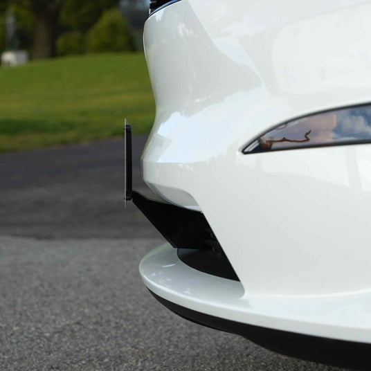 Close-up of a white cars rear bumper with a black accent piece, parked on a paved surface with grass and trees in the background.