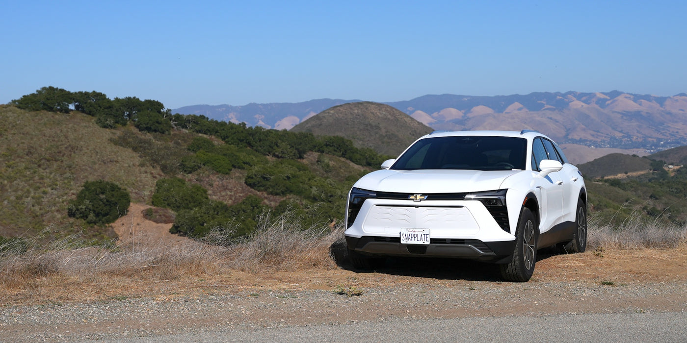 A white electric SUV is parked on a rural roadside, with rolling hills and a clear blue sky in the background.