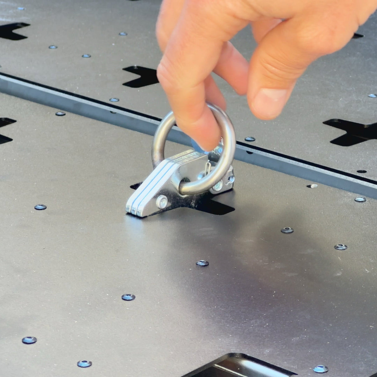 A hand lifts the metal ring latch of the Thunderbolt Adventure Supply R1T Bed Glider, a secure, screw-mounted accessory designed for versatile truck bed storage and camping solutions.