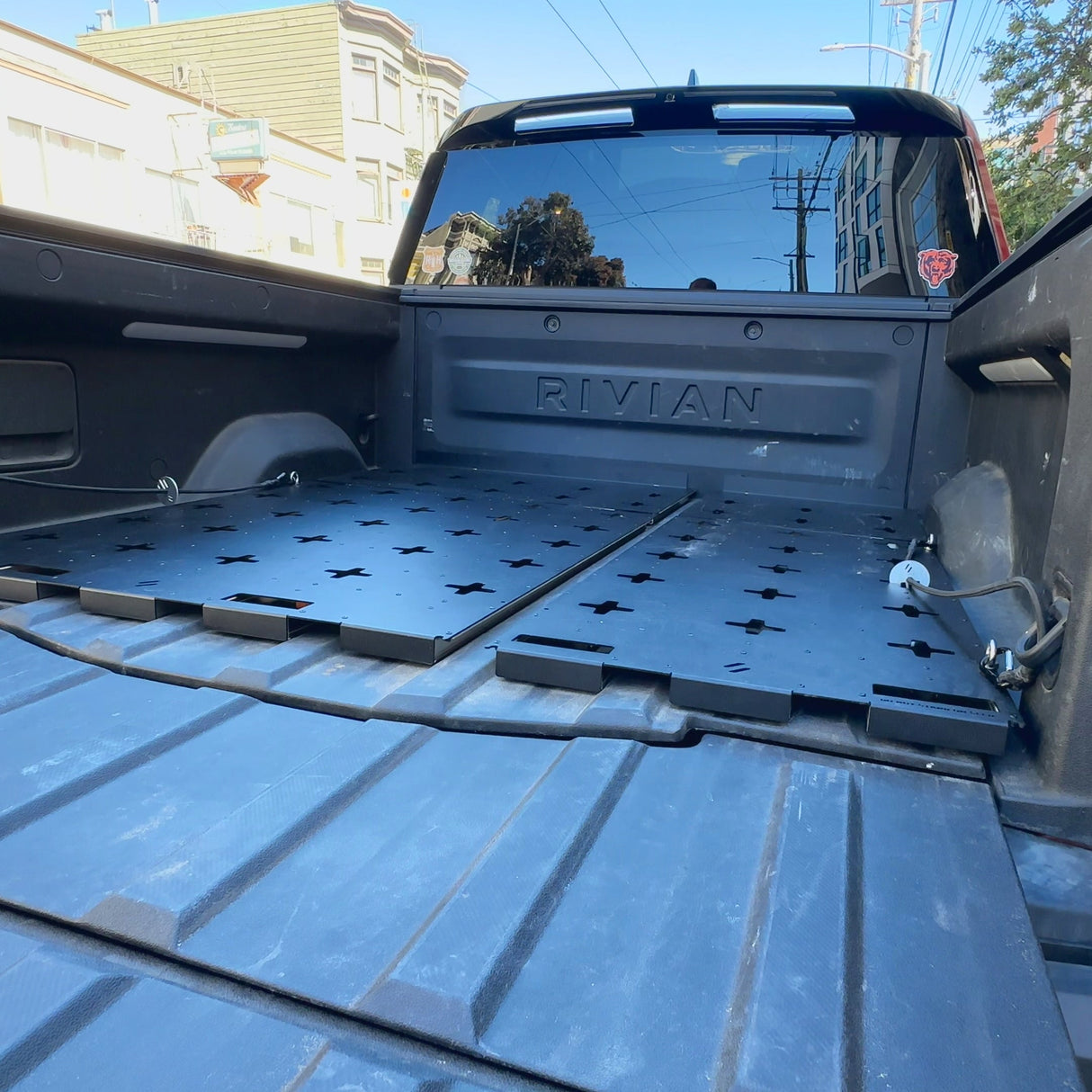 A Rivian pickup’s truck bed features the Thunderbolt Adventure Supply R1T Bed Glider, a modular cargo platform—ideal for boosting storage or organizing camping gear, shown in an urban outdoor setting.