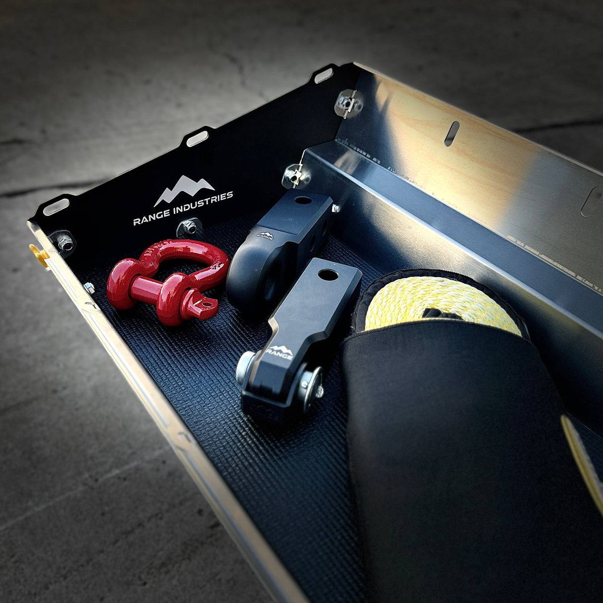 The Gear Tunnel Slide 'LITE' by Range Industries is a versatile Rivian R1T storage solution, holding two red shackles, two black tow hitch receivers, a yellow rope, and a branded black pouch—all in a sturdy metal box.