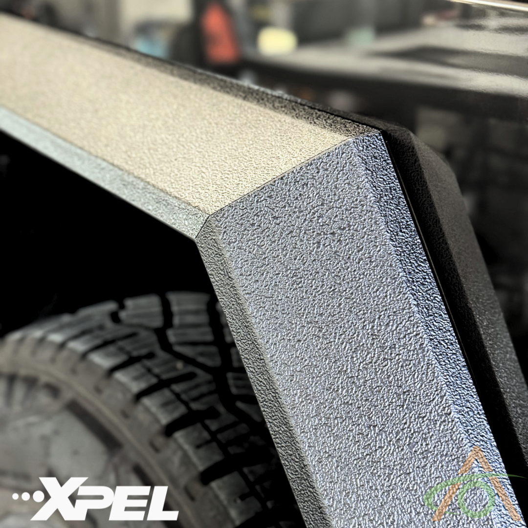 A close-up shows the rough texture of Ultra-Tough Xpel Armor Fender Flares from Abstract Ocean on a Cybertruck wheel arch, featuring parts of the tire and branding logos. The plastic trim protection enhances durability.