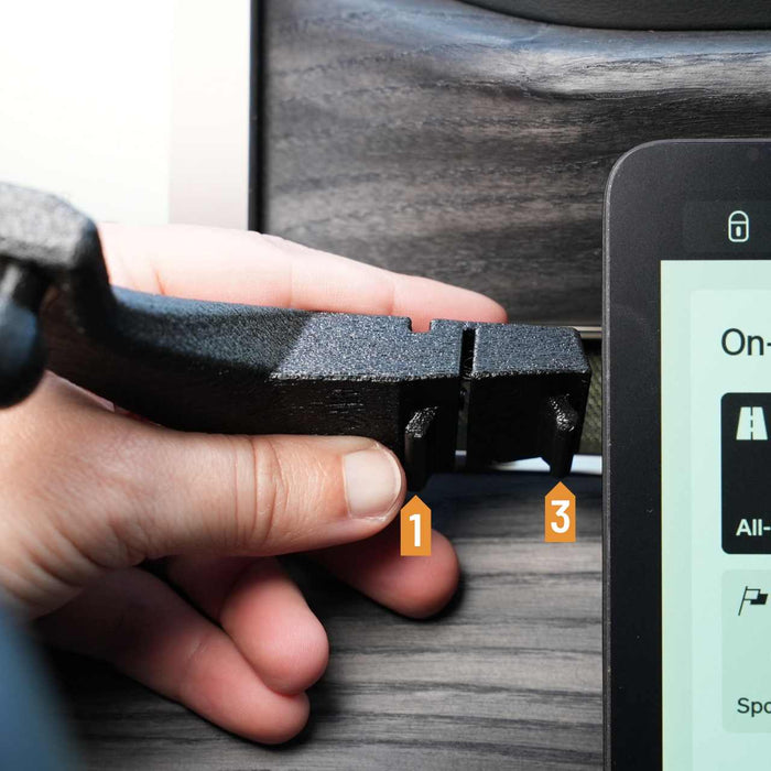 A hand holding a black plastic clip alongside a digital display on a wood surface, with numbered markers showing dimensions.