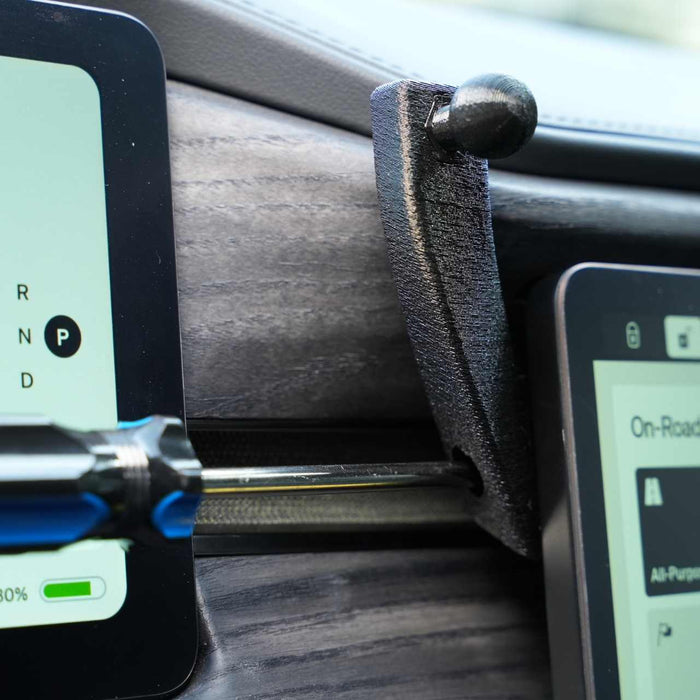 A screwdriver is inserted between two touchscreens in a vehicle dashboard, set against a wood grain panel.