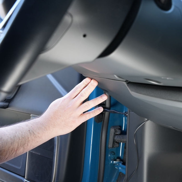 A persons hand reaching under the steering column of a vehicles dashboard, close to a blue panel and a speaker.