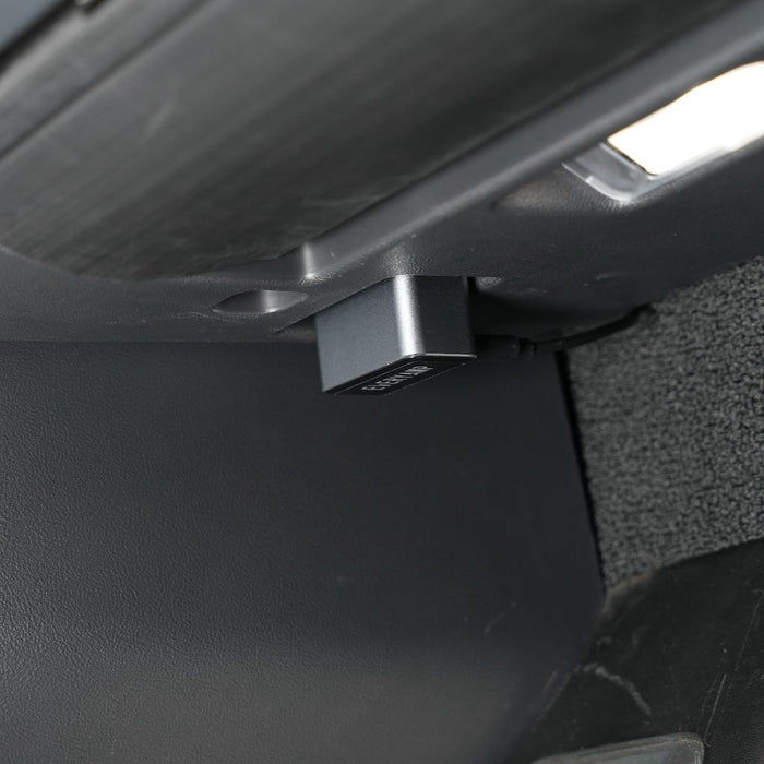 A small black device with a label is plugged into a socket below a dashboard inside a vehicle. The cars interior is partially visible, including textured and smooth surfaces.