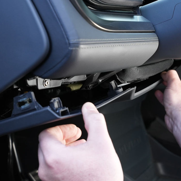 Hands are removing a cars glove compartment panel, exposing the interior components beneath the dashboard.