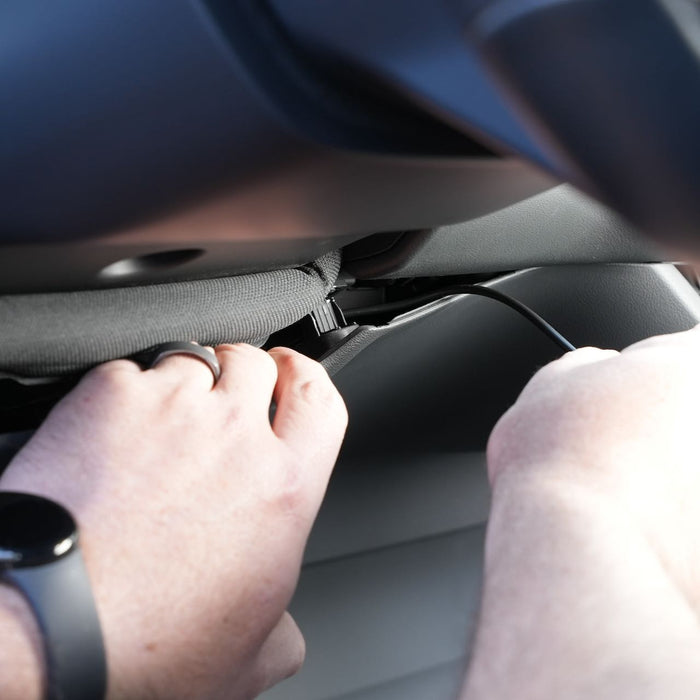 Hands adjusting cables under a car dashboard, wearing a wristwatch and a ring.