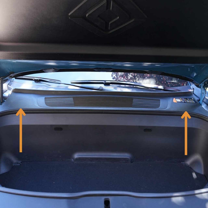An open car hood reveals the storage compartment beneath. Two orange arrows point upward, likely indicating features or components of interest inside the compartment.