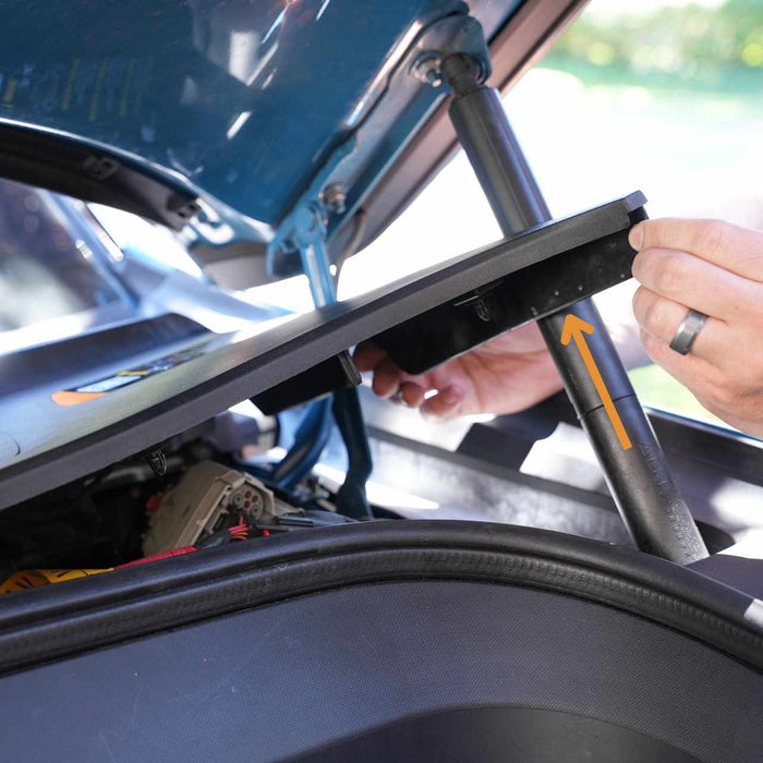 Hands adjusting a panel inside an open car trunk, with a visible orange arrow indicating direction.