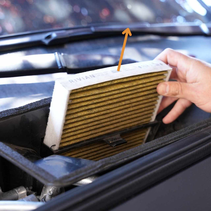 A hand pulls out an air filter from a vehicle compartment, with an arrow pointing towards the filter.
