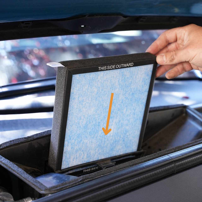Hand inserting a blue air filter labeled THIS SIDE OUTWARD into a cars cabin air filter compartment under the hood.