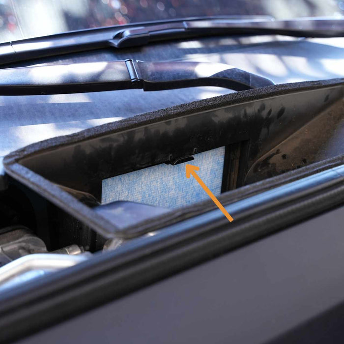 Car engine compartment with an exposed air filter highlighted by an orange arrow. The filter is visible through an open panel near the windshield wipers.