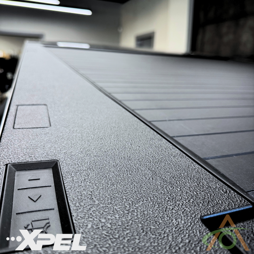 Close-up of a black textured surface with grooves, featuring button-like rectangles on the side. Logos in the corners read XPEL and display an abstract design, showcasing Abstract Oceans Ultra-Tough Xpel Armor Bed Protection for Cybertruck, ensuring ultimate durability.