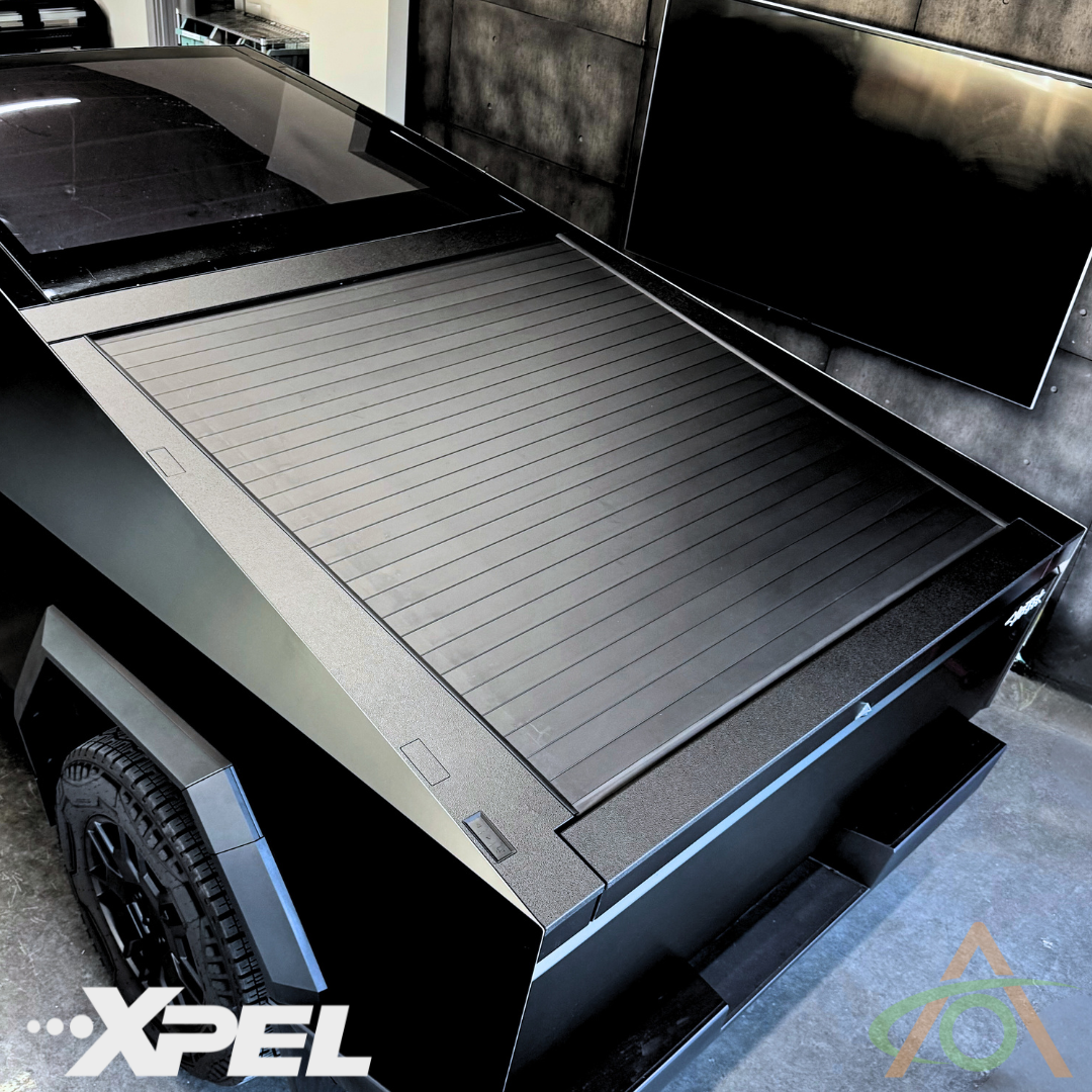 A futuristic black truck with a retractable cover is parked inside a modern garage, featuring Abstract Oceans Ultra-Tough Xpel Armor for Cybertruck protection. The XPEL logo gleams in the foreground.