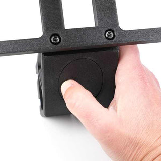 A hand presses a button on a black rectangular device attached to a metal frame with screws.