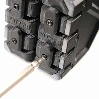 Close-up of a black mechanical device with multiple components held together by screws. A screwdriver is positioned in one screw, suggesting an adjustment or assembly in progress.