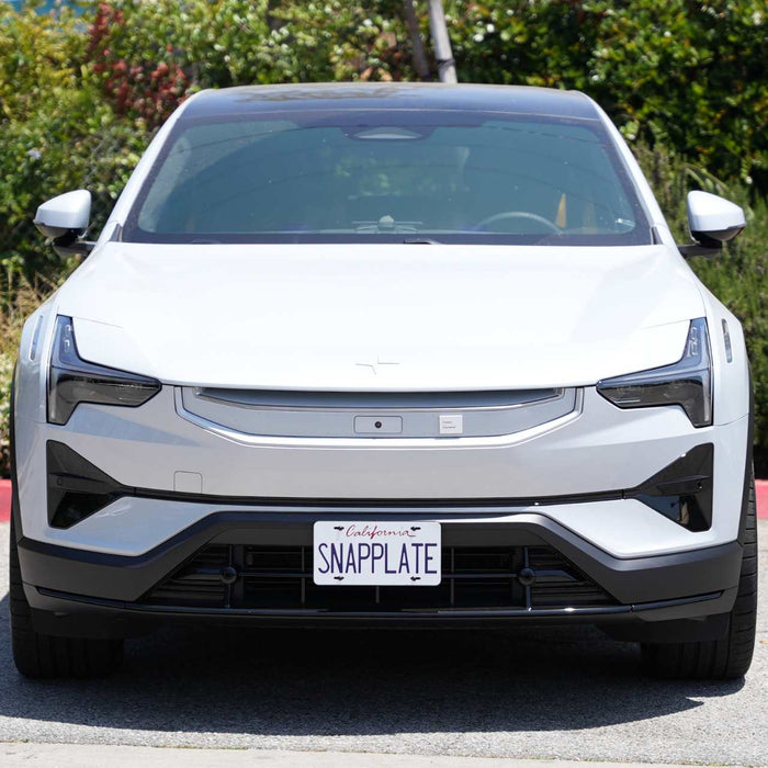 Front view of a white electric car with a SNAPPLATE license plate, parked on a street with greenery in the background.