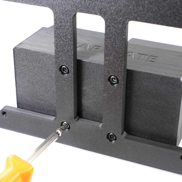A close-up of a screwdriver tightening a screw into a black metal bracket with four screws already installed.