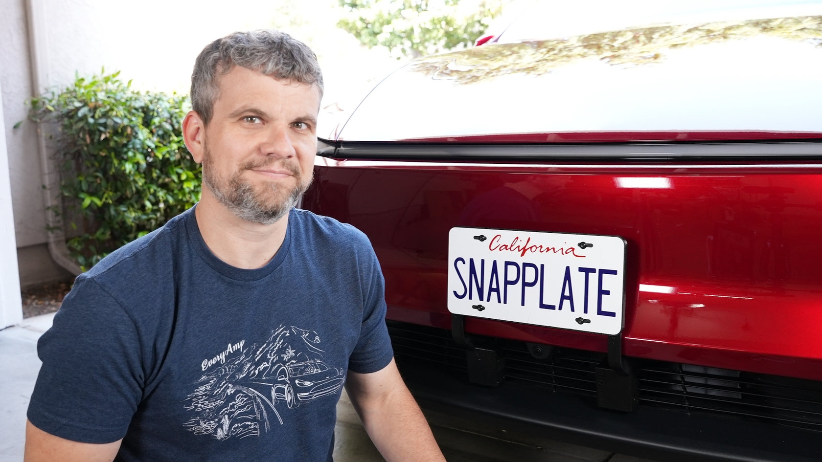 A man kneels next to a red car with a California license plate that reads SNAPPLATE in blue letters.