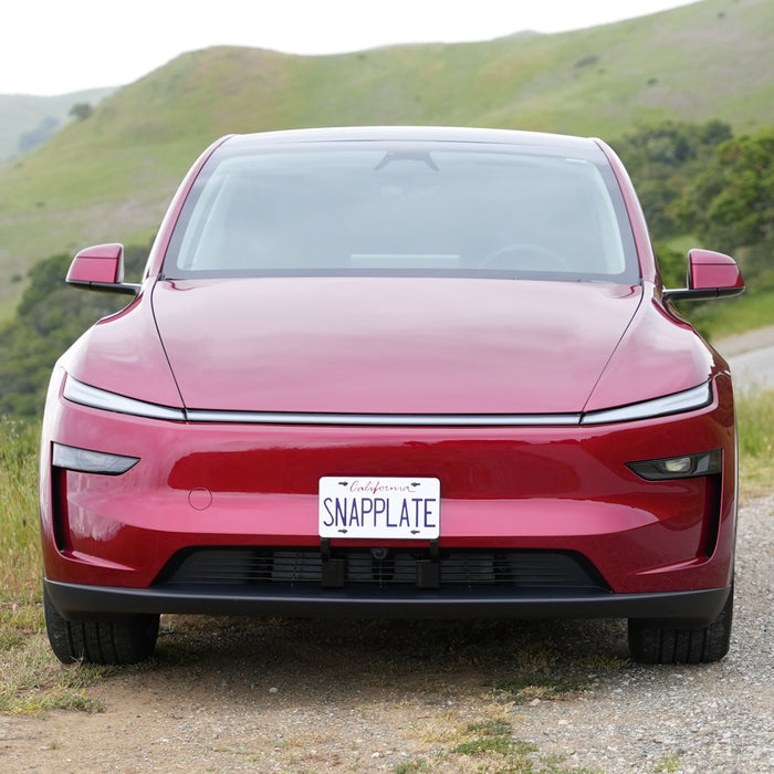 A red Tesla Model Y with sleek headlights is parked on a rural road, featuring a California plate mounted with the EveryAmp Tesla Model Y SnapPlate Front License Plate Holder. Green hills create a scenic backdrop.