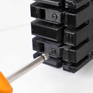 A screwdriver with an orange handle is tightening a screw into a stack of black plastic or metal components with multiple screws visible.