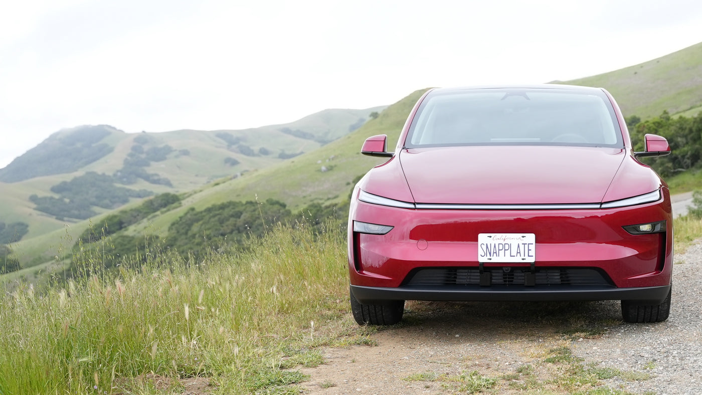 A red electric car with a SNAPPLATE license plate is parked on a roadside with green hills in the background.