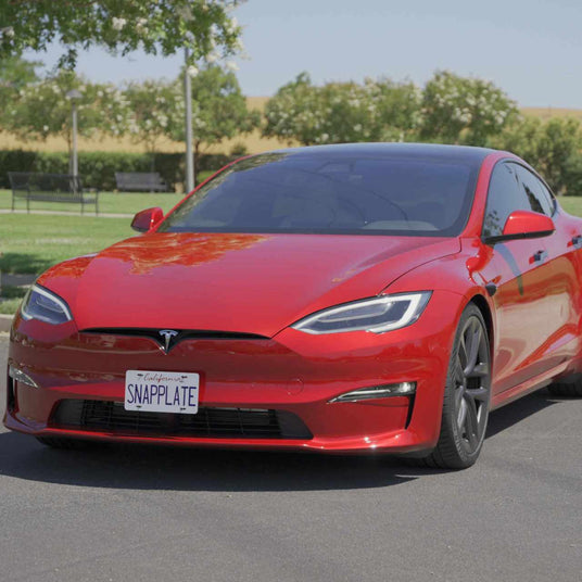 Red Tesla car parked on a street, featuring a license plate that reads SNAPPLATE. Trees and a field are visible in the background.