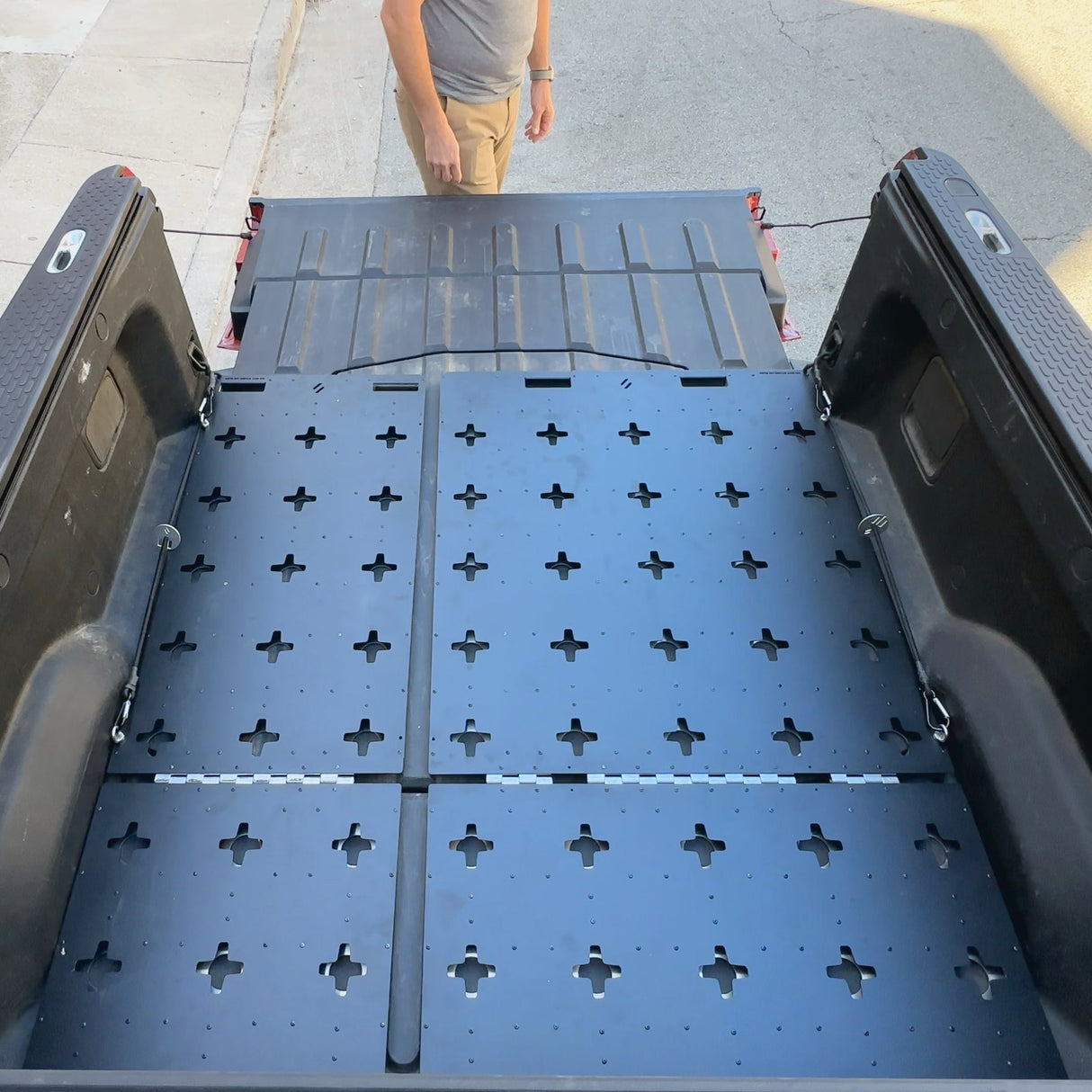 A person stands near the open tailgate of a pickup truck equipped with the Thunderbolt Adventure Supply R1T Bed Glider, showcasing its black, perforated bed cover and organized storage system in the bed.
