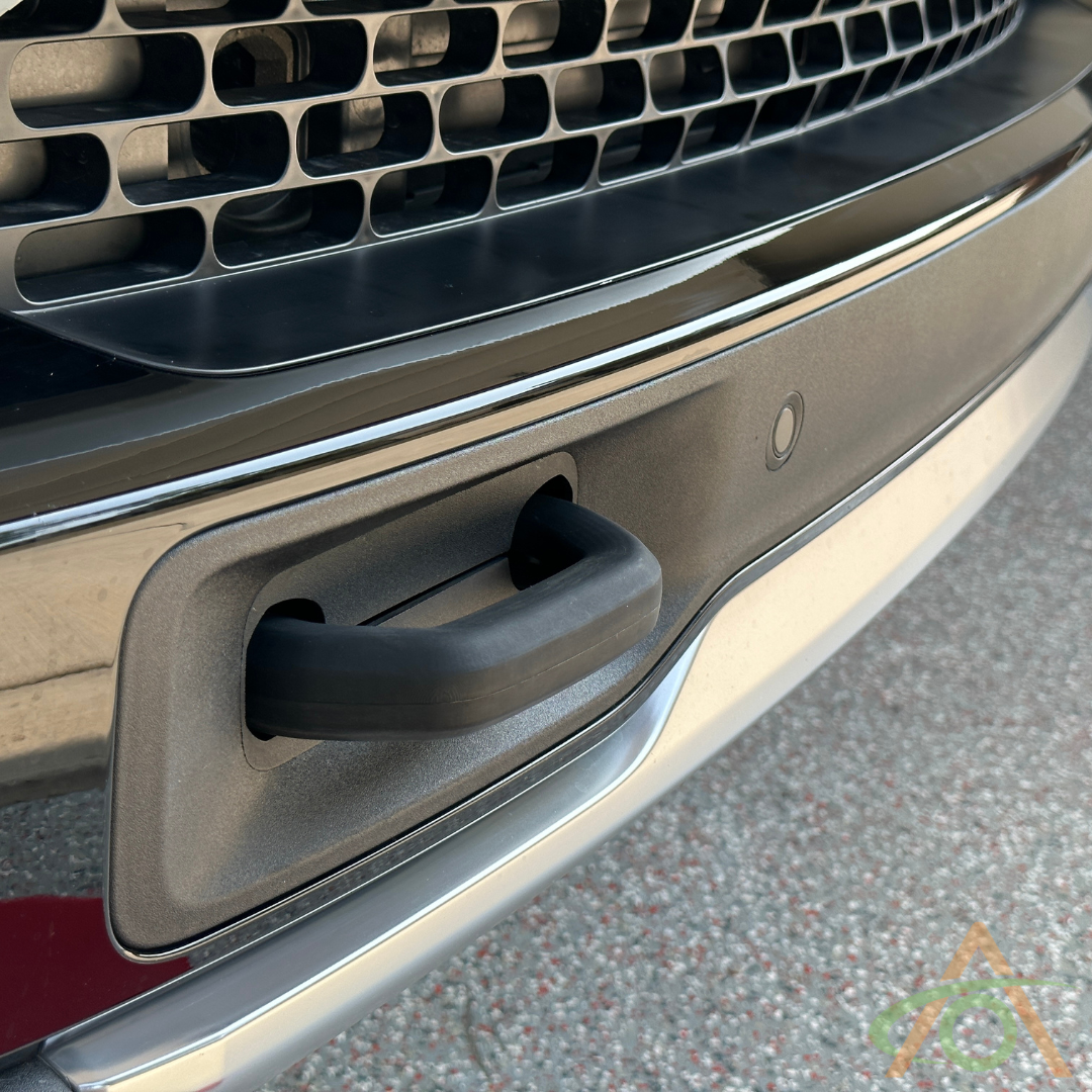 Close-up of a vehicles front bumper showing a visible tow hook with textured grille above, enhanced by sleek Rivian accessories, including black Silicone Hook Covers by Abstract Ocean for a perfect touch.