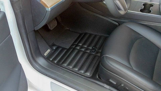 Car interior showing a drivers seat and floor mat with a textured design. The steering wheel and pedals are partially visible.