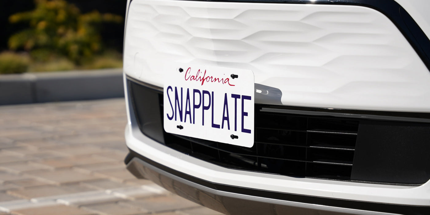 Close-up of a front car bumper with a California license plate reading SNAPPLATE. The plate is white with red and blue letters, mounted on a white vehicle.