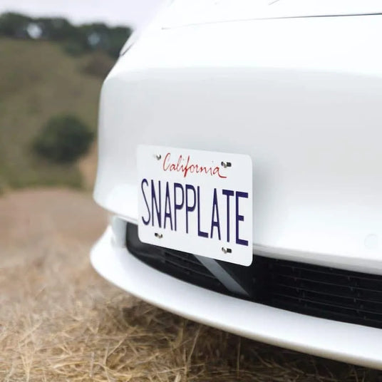 Close-up of a white car with a license plate reading SNAPPLATE and California in red script, parked on dry grass with blurred trees and hills in the background.