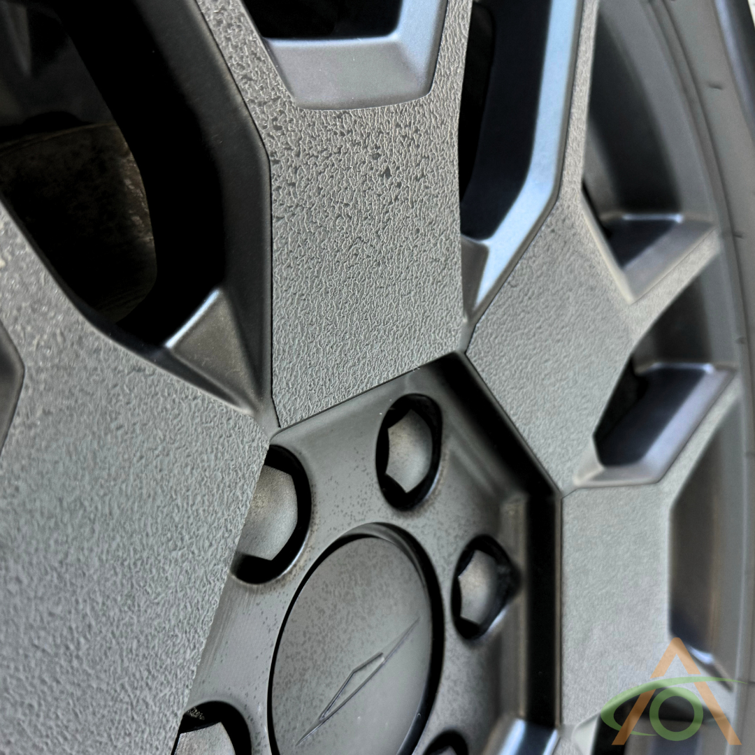 Close-up of a silver alloy wheel with a textured surface and geometric design, featuring hexagonal and circular elements. Highlighted by Abstract Oceans Xpel Armor All-Terrain Wheel Decals for Cybertruck, a small colorful logo is visible in the lower right corner.