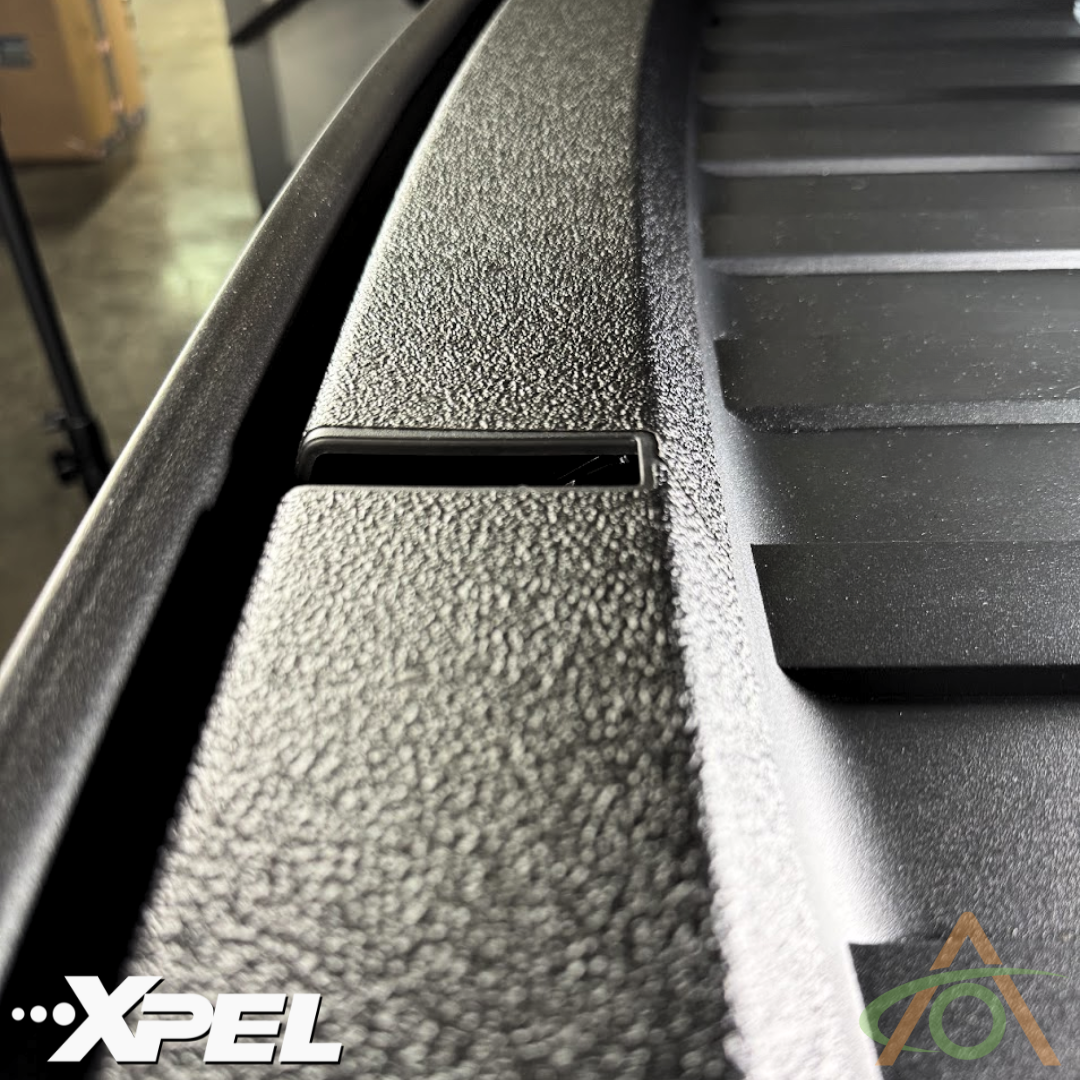 Close-up of a textured black surface with parallel ridges and a small rectangular cutout, enhanced by Abstract Oceans Ultra-Tough Xpel Armor Frunk Protection for Cybertruck. The Xpel logo is visible in the corner, indicating its role in safeguarding against wear.