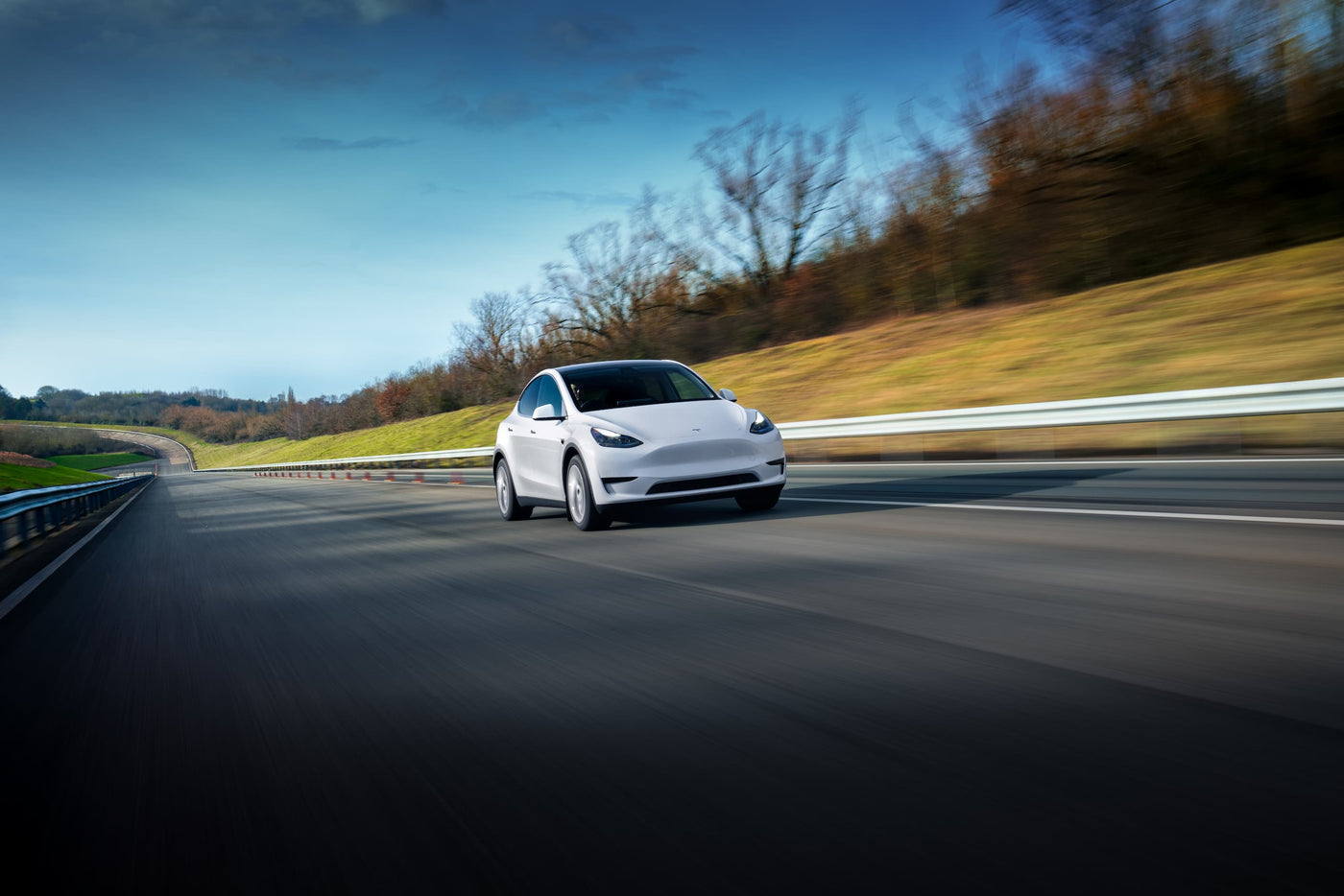 A white car is driving on a highway surrounded by grassy hills and leafless trees under a clear blue sky.