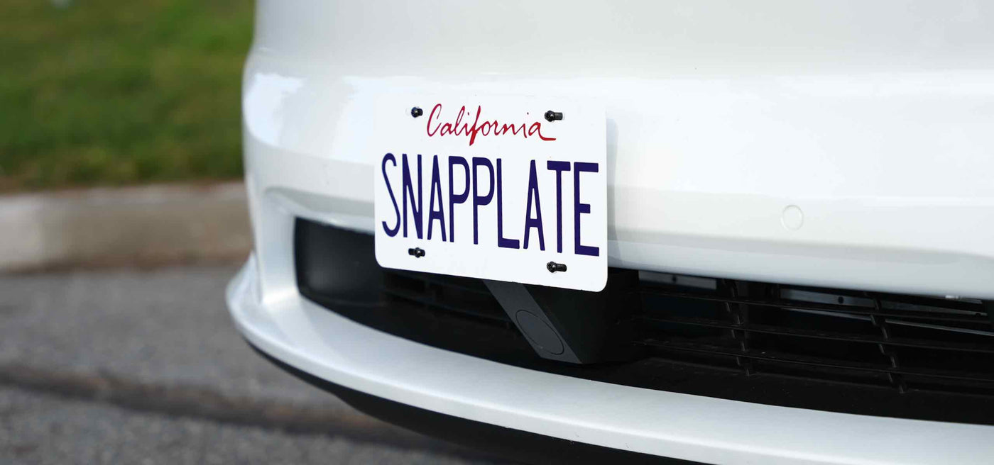 White car with a California license plate reading SNAPPLATE on the front bumper, parked on a paved road.