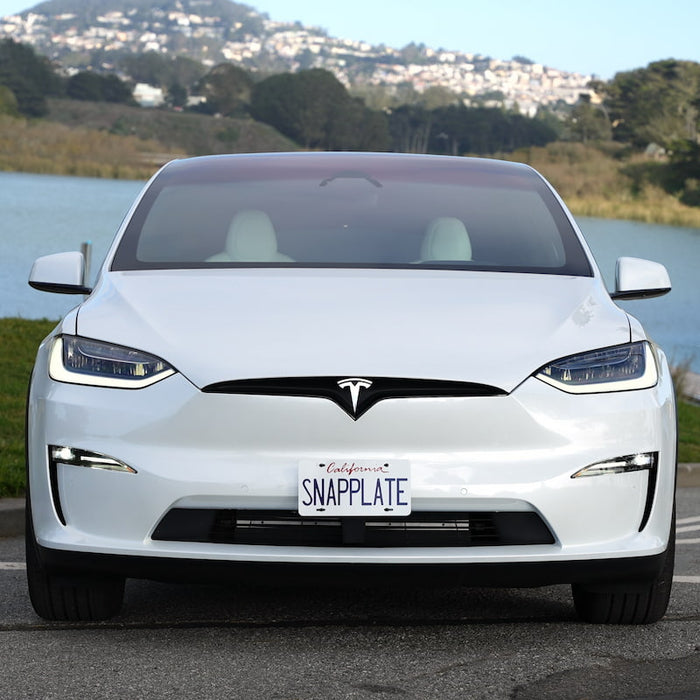 Front view of a white Tesla with a SNAPPLATE license plate, parked near a body of water with hills in the background.