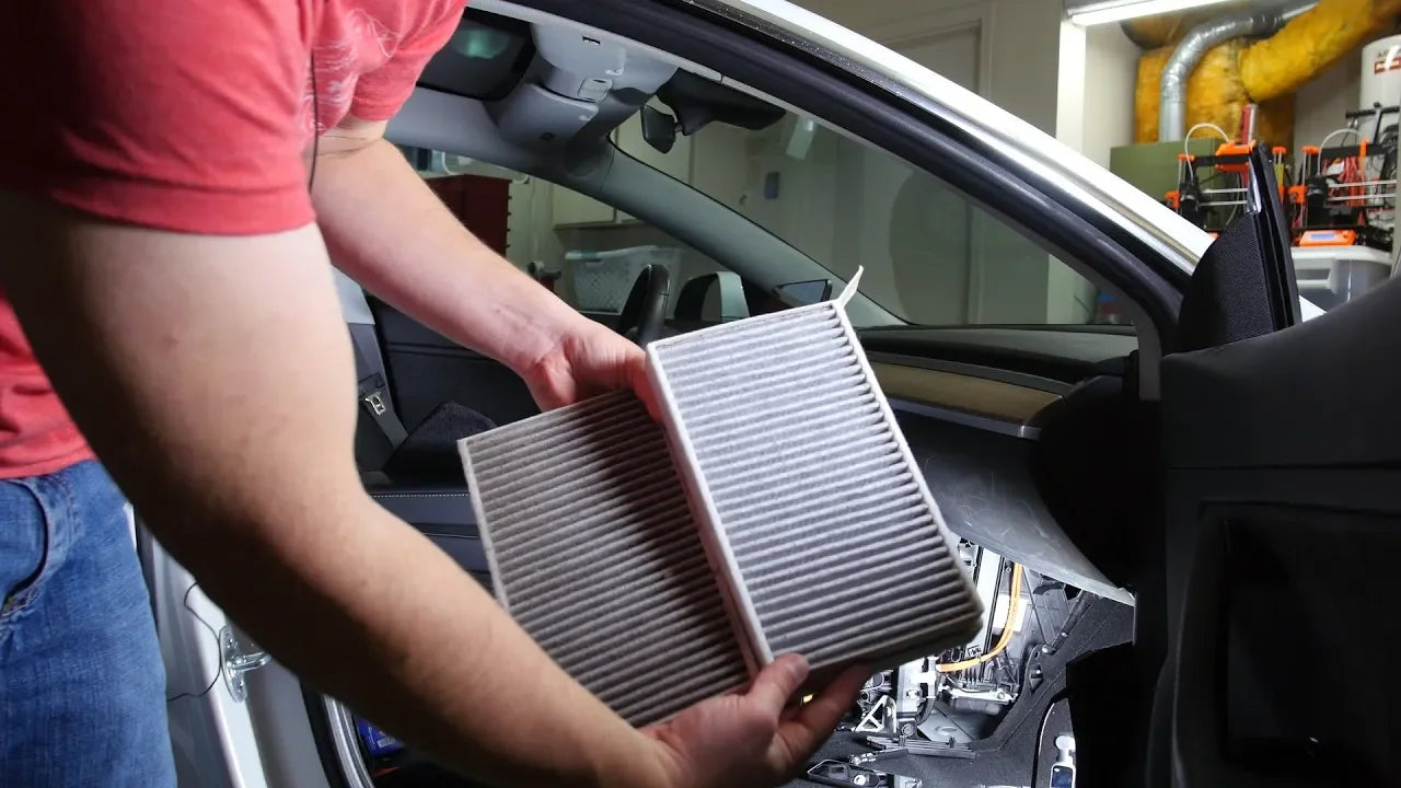 Person replacing a car cabin air filter inside a vehicle. The glove box is open, and tools are visible on the floor.